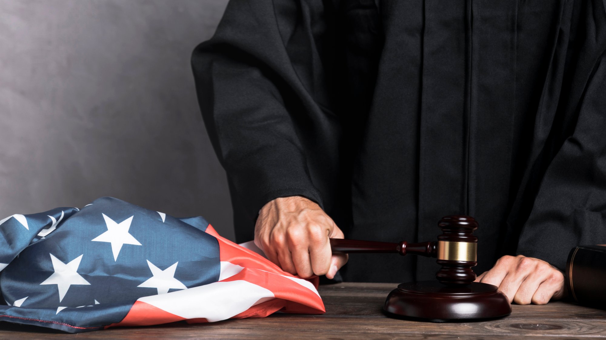 A judge in a black robe holds a gavel next to a crumpled American flag on a wooden table. The image conveys authority and justice. A judge in a black robe holds a gavel next to a crumpled American flag on a wooden table. The image conveys authority and justice.