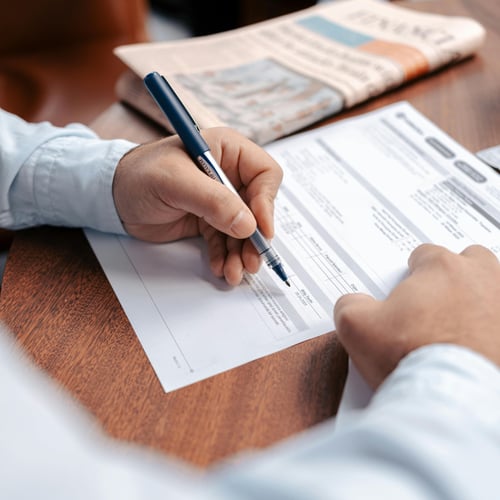 A person in a light blue shirt reviews financial documents with a pen on a wooden table. A newspaper is slightly blurred in the background.