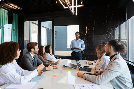 A diverse group of colleagues in a modern conference room watches a colleague standing and presenting confidently, fostering collaboration and engagement.