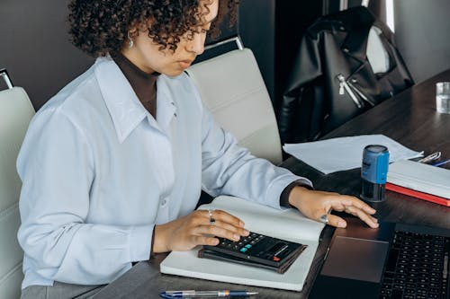 A woman working in a regulatory industry using a calculator and laptop while reviewing financial documents at an office desk.
