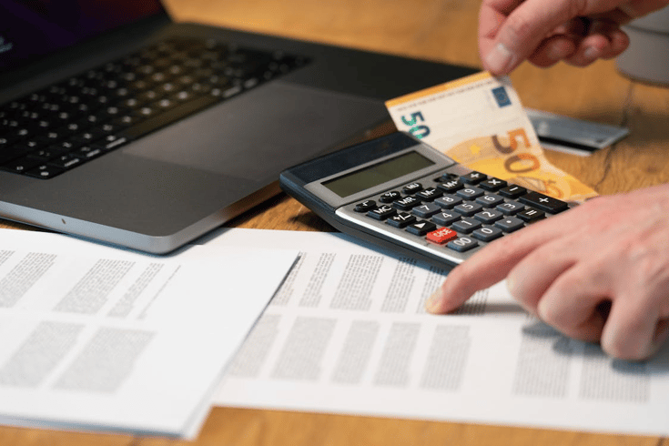 A person using a calculator and reviewing printed documents beside a laptop, calculating implementation and maintenance costs for regulatory compliance software.